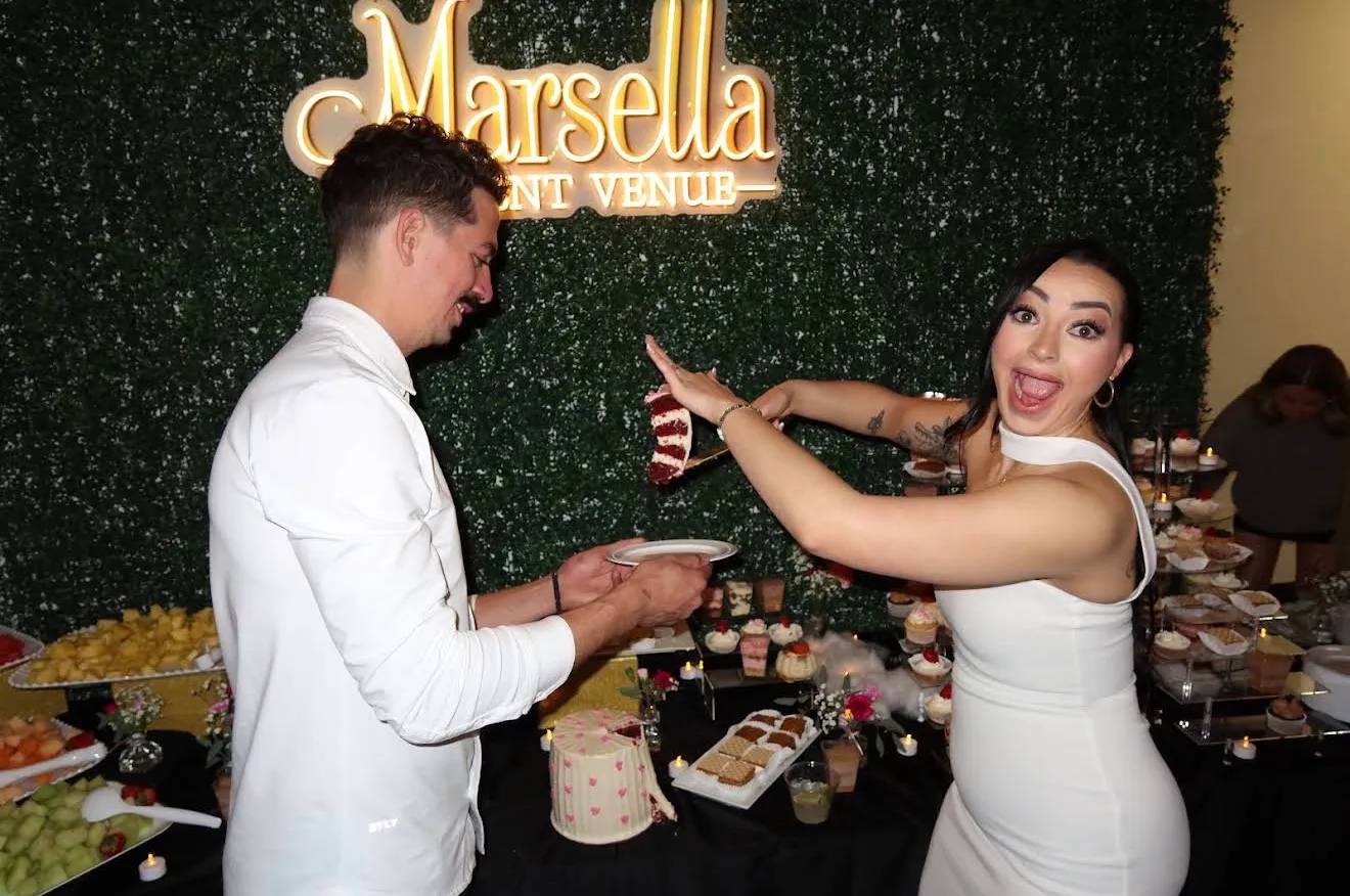 A couple cutting their cake in front of the Marsella sign on the greenery wall