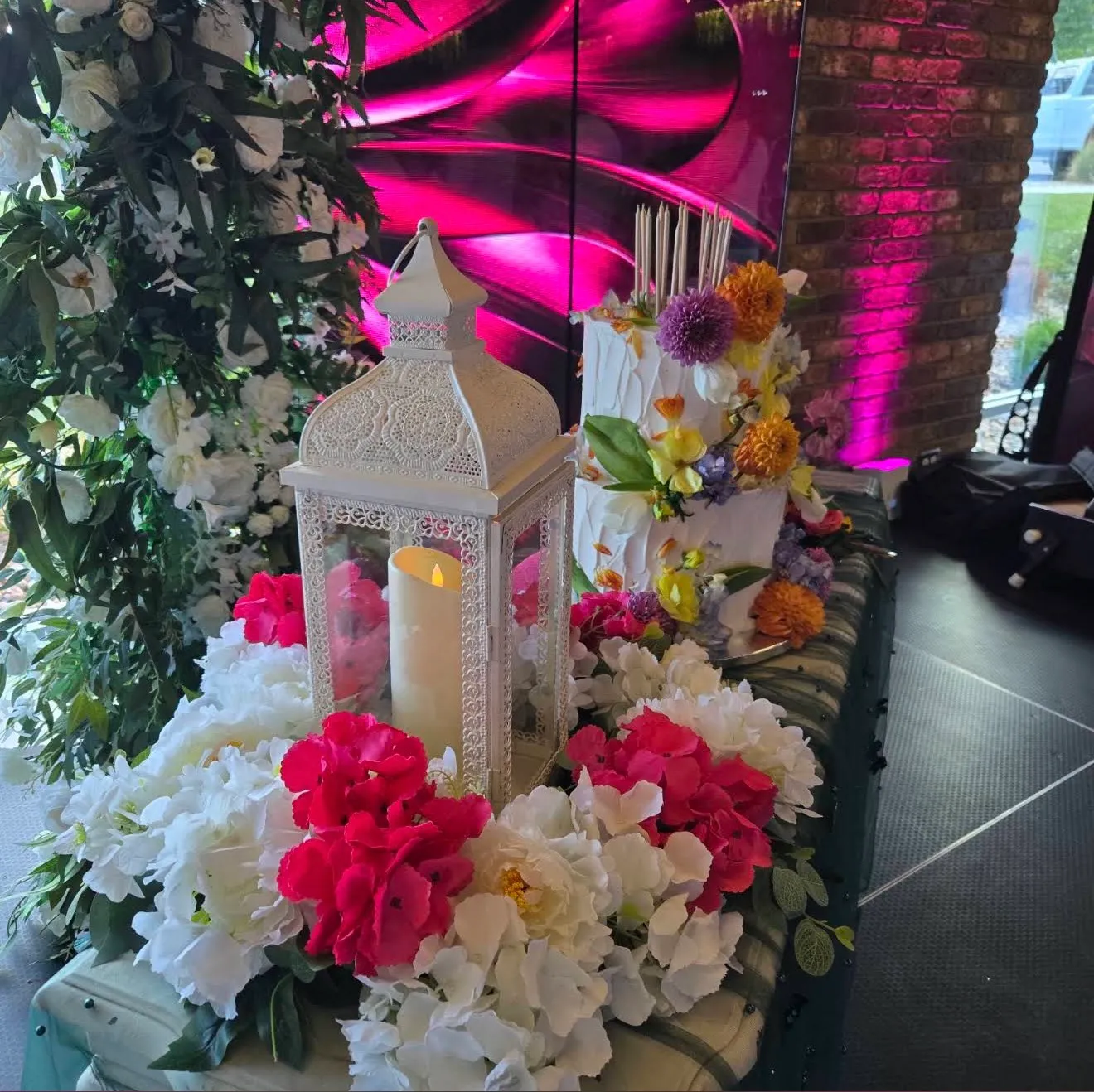 A flower-topped cake beside a white lantern, LED screens glowing magenta