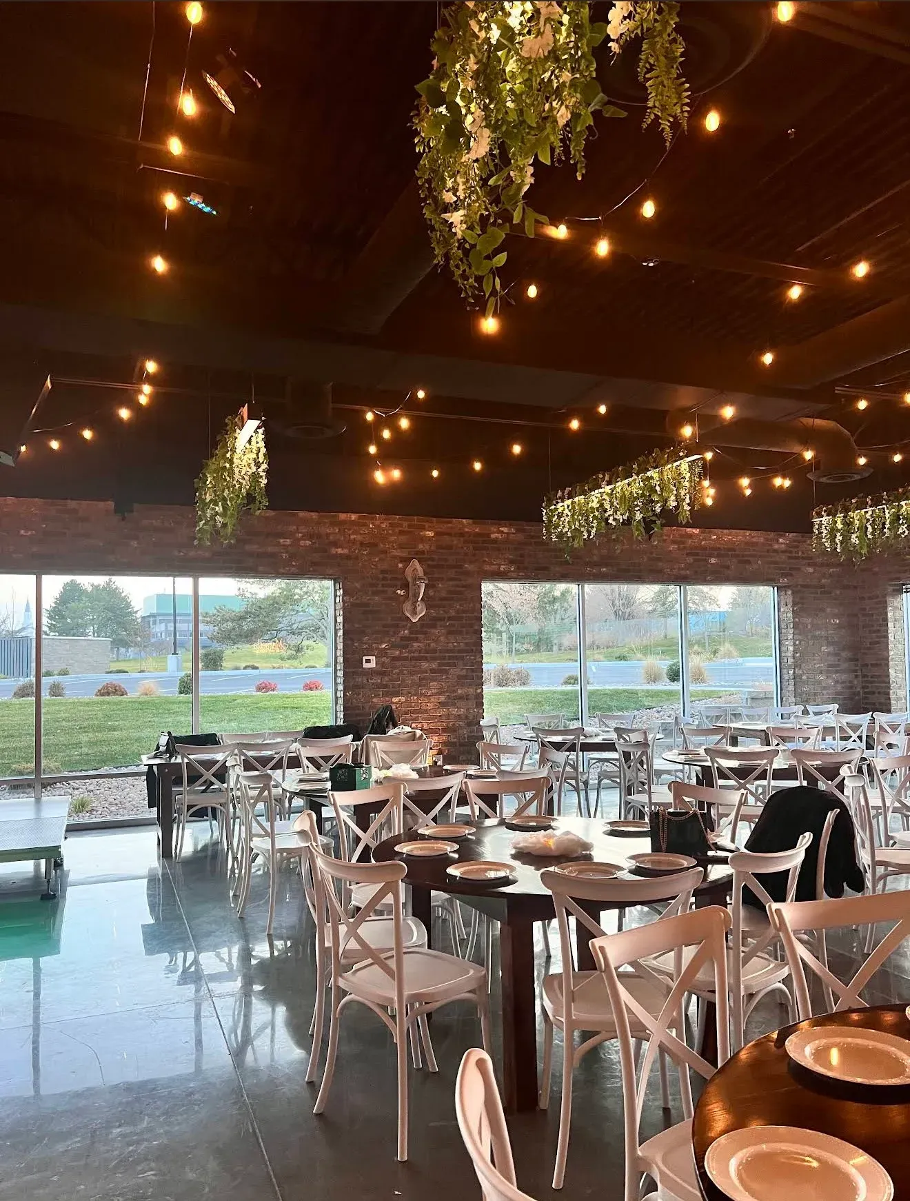 The room by day: round wood tables, white chairs, string lights and tall windows