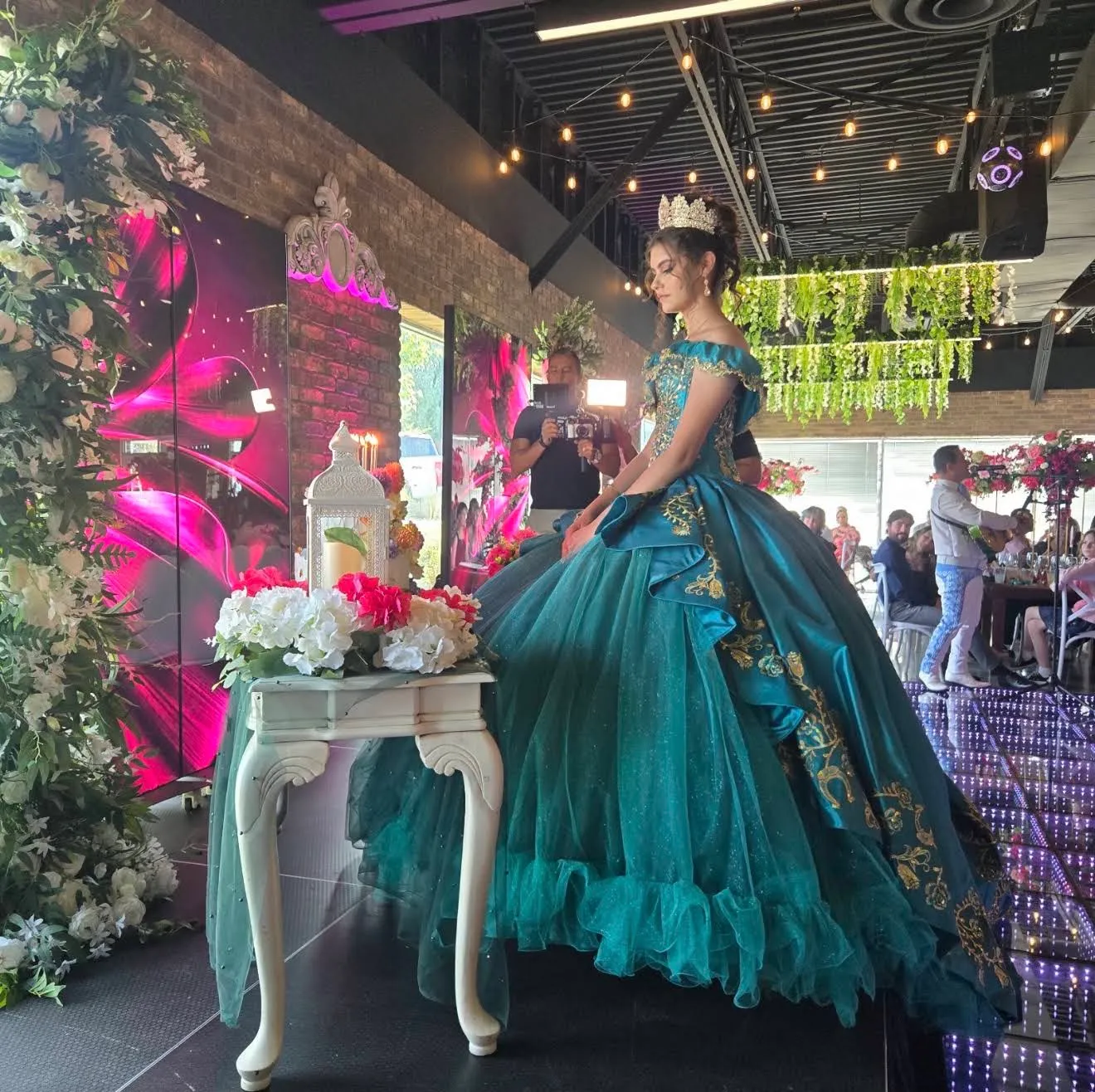 A quinceañera in an emerald gown and crown on the stage, mariachi playing behind