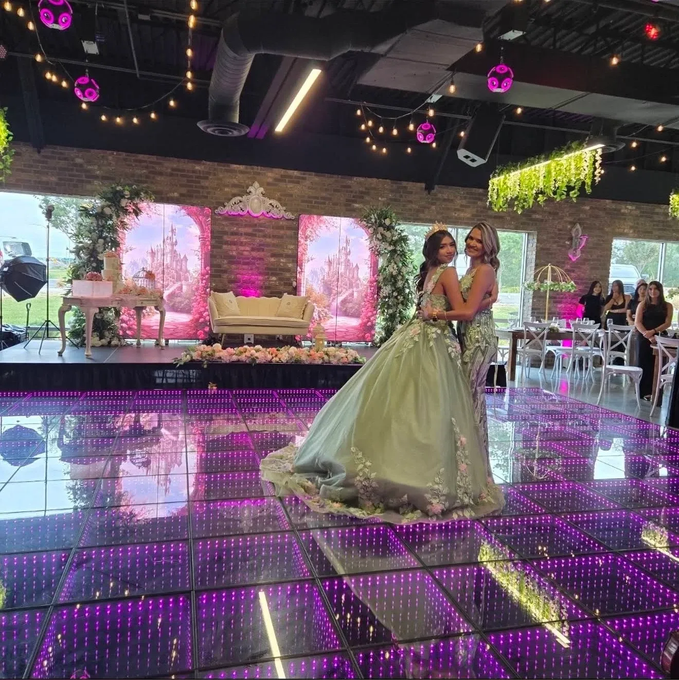 A quinceañera in a sage gown hugging her mom on the purple LED dance floor, castle scenes on the LED screens behind them